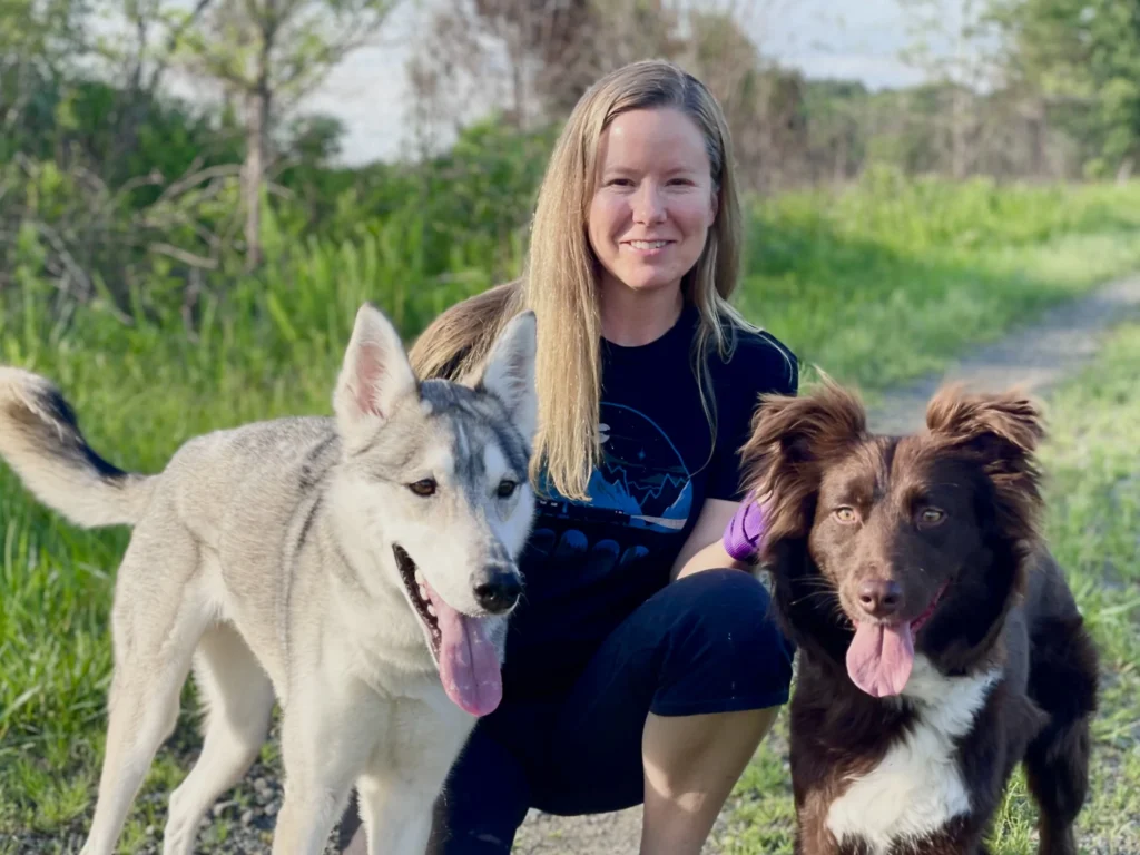 A smiling woman with long blonde hair is kneeling on a gravel path outdoors, flanked by her two dogs. On the left stands a lean Husky and German Shepherd mix with a light grey and white coat, erect ears, and its tongue happily lolling out. On the right stands a fluffy, dark brown and white Australian Shepherd with perky ears, also panting with its tongue out. The background features a sunny, grassy field lined with green trees and bushes.